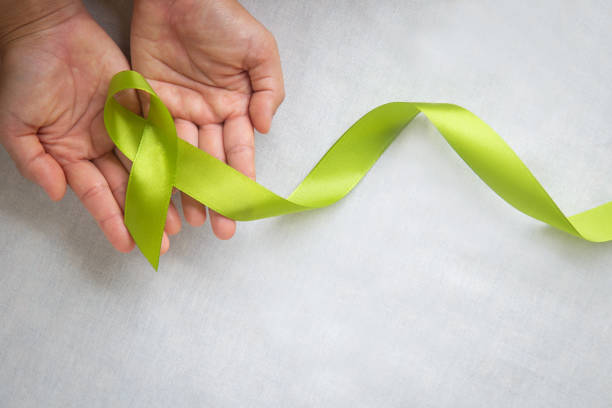 Hands holding light lime green ribbon on white fabric background with copy space. World Mental Health Day and Lymphoma Awareness symbol. Healthcare medical and insurance concept.